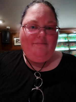 a white woman with dark hair tied back and glasses, wearing a black top and necklace with three large rings
