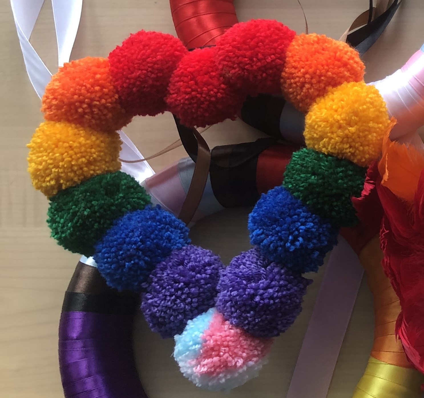 Wreath in a heart-shape made from yarn pom-poms in rainbow colours with trans colours