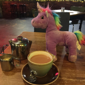 a purple plush unicorn with a rainbow mane and tail sitting on a table next to a cup of tea